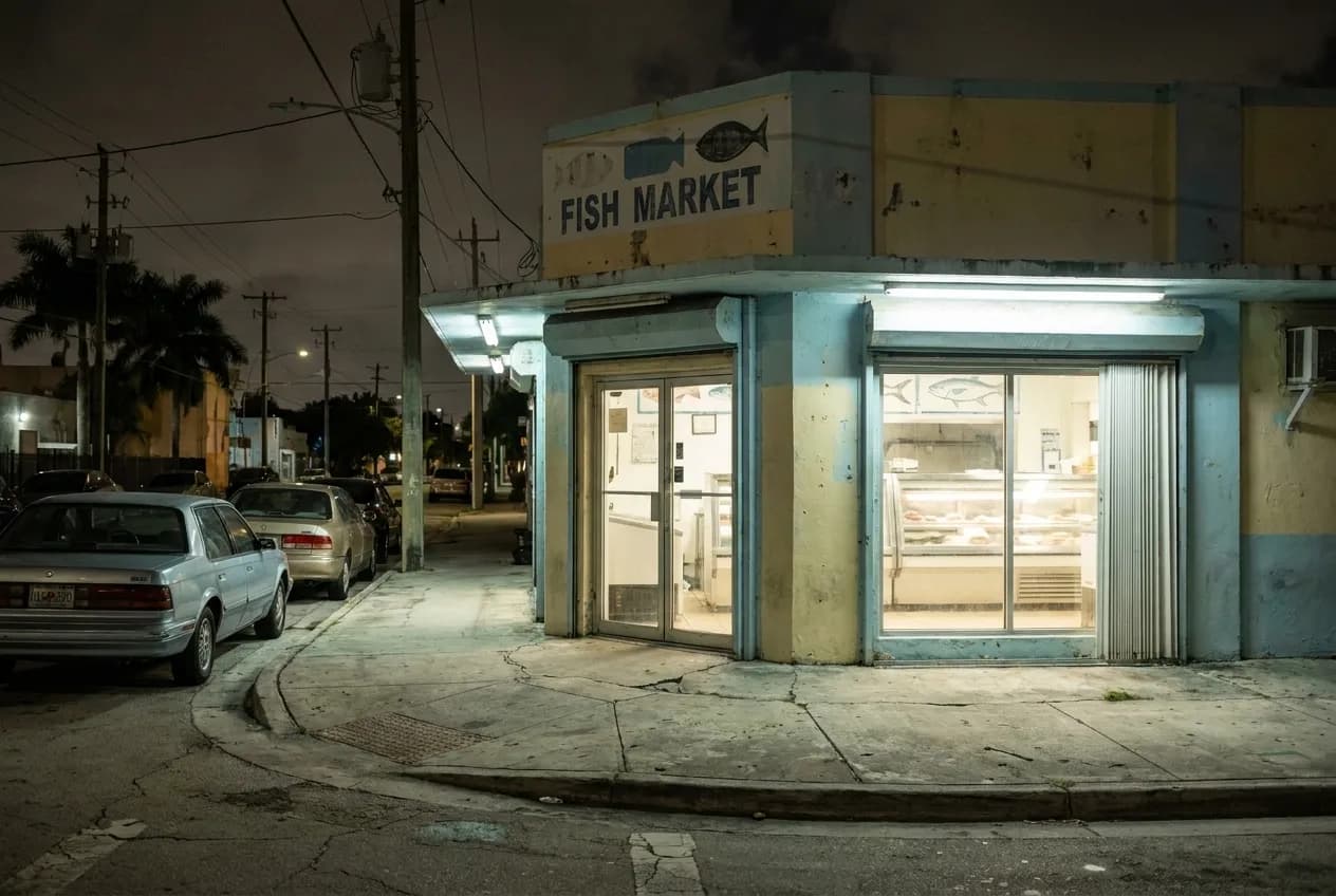 Evening view of a modest concrete fish market storefront in Allapattah with fluorescent-lit display cases visible through open doors and empty metal tables outside.