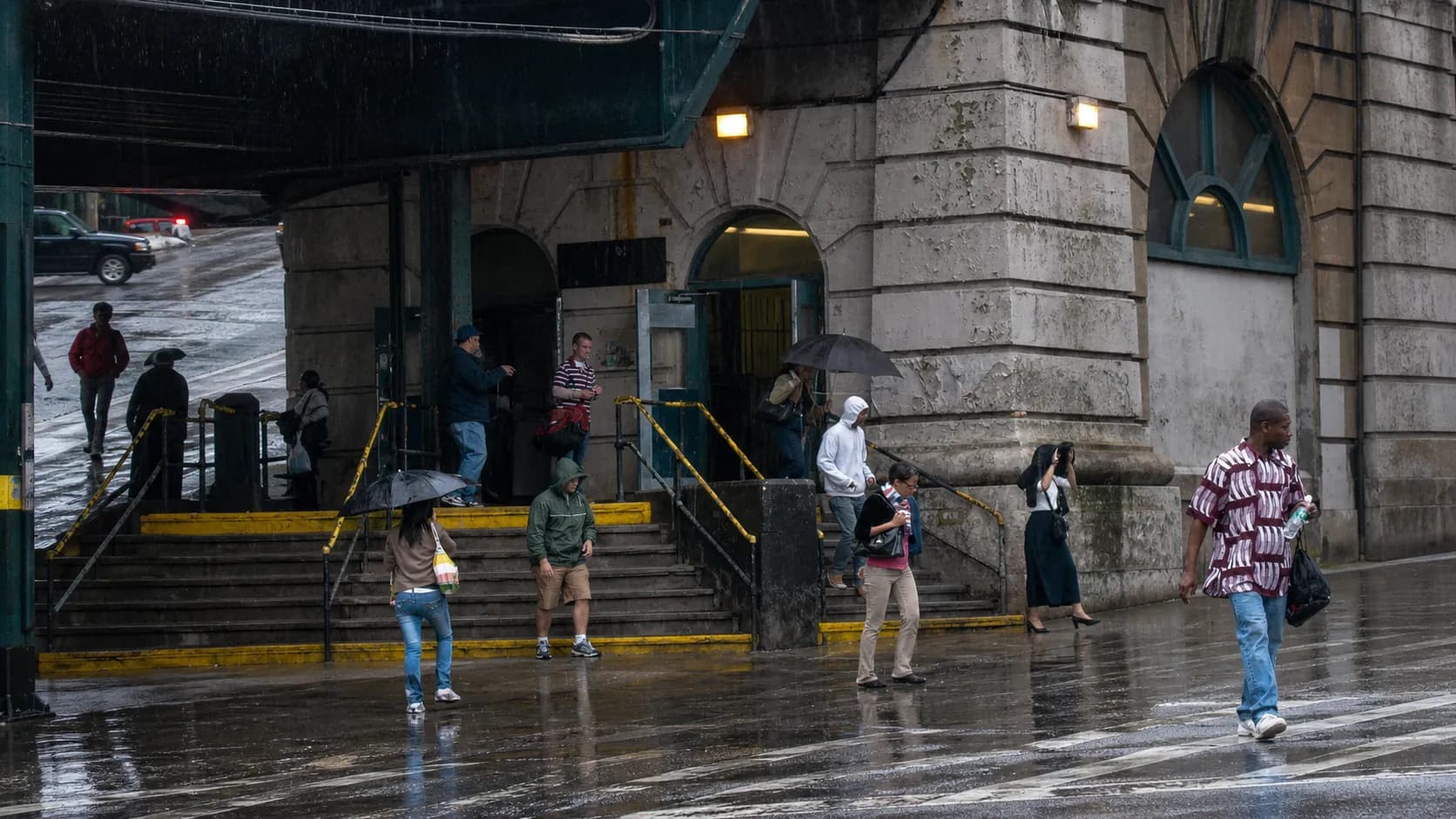 Commuters with umbrellas walk near a wet New York City subway entrance.