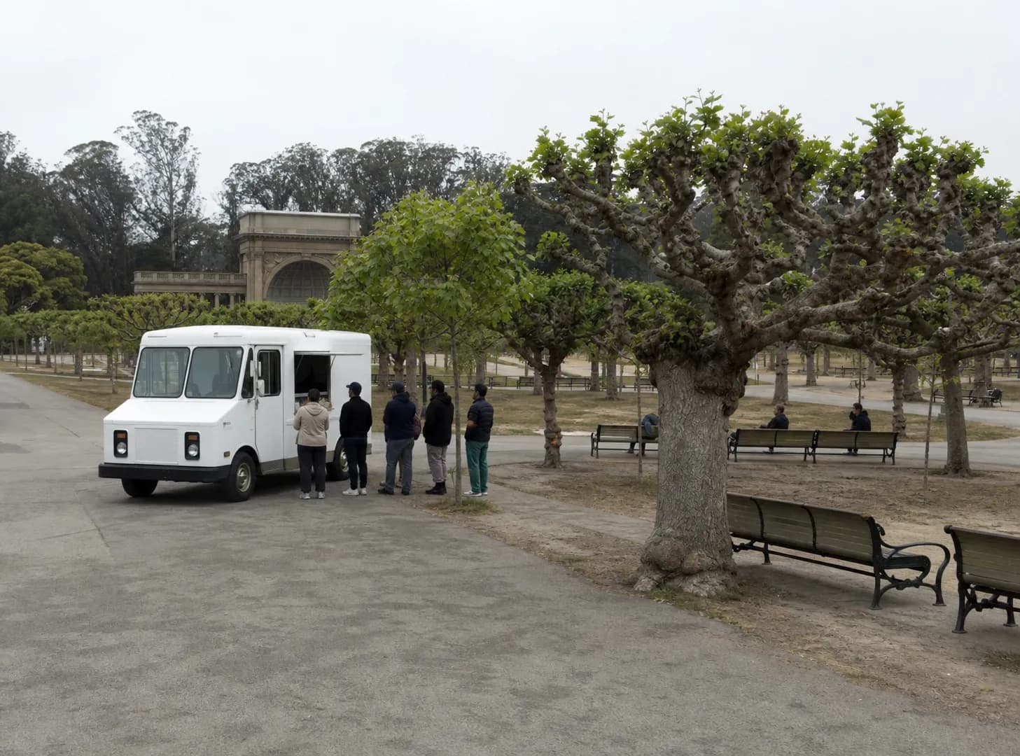 A plain white ice cream truck parked at the curb of the tree-lined Music Concourse in Golden Gate Park with a short line of people waiting, under flat overcast San Francisco light