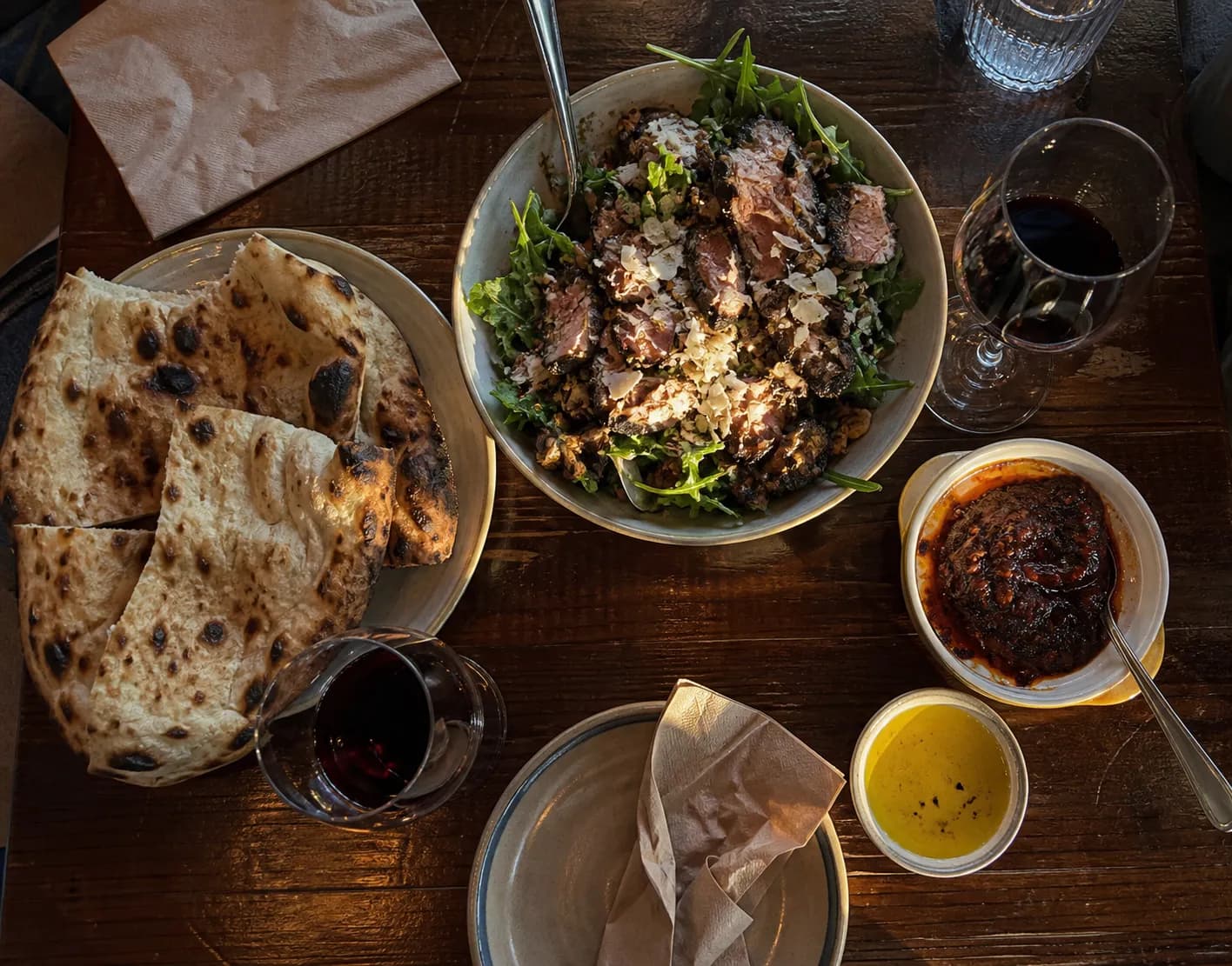 Overhead view of a shared dinner table in a casual San Francisco taverna with charred flatbread, a roasted-meat salad bowl and two glasses of red wine