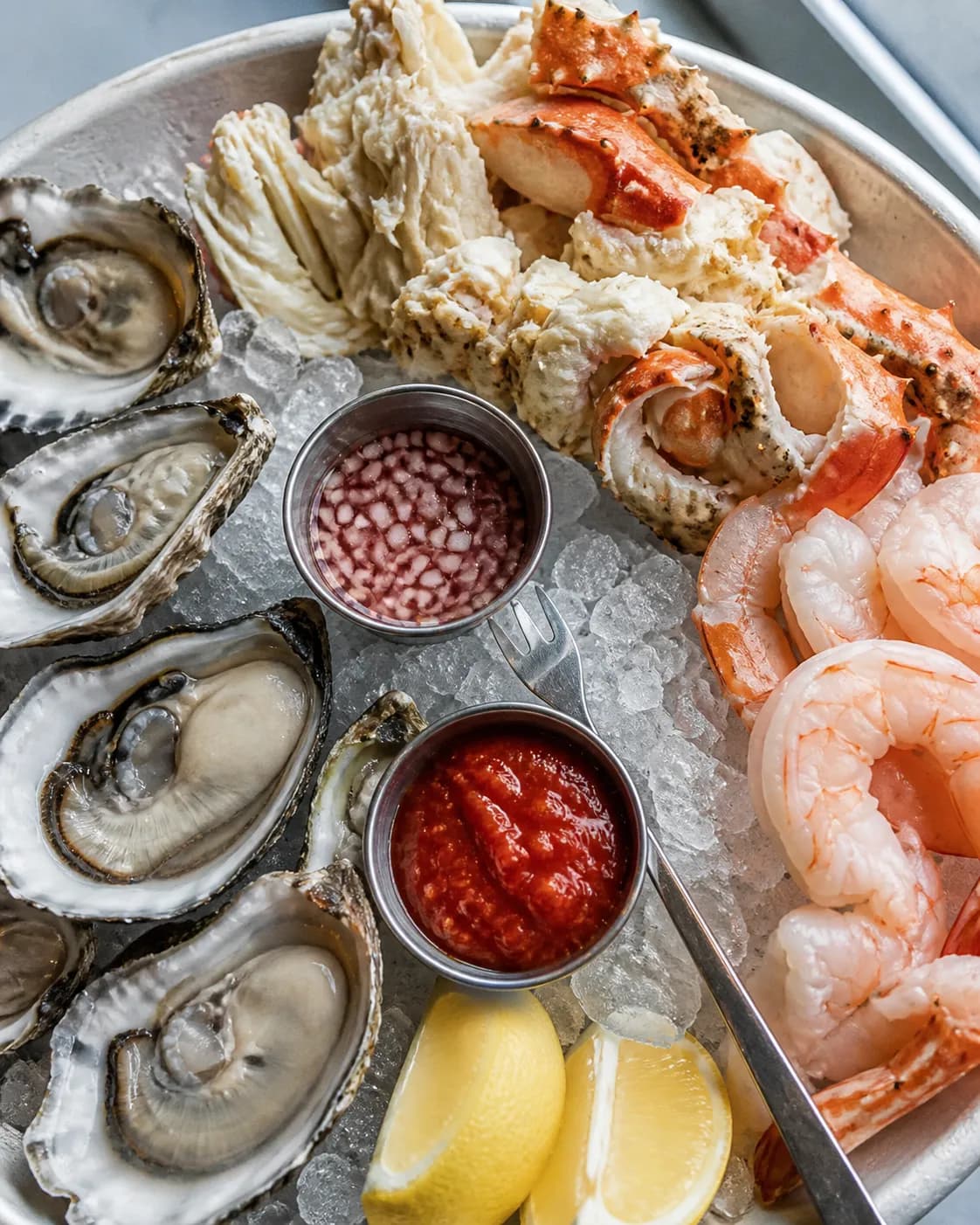 Overhead view of a chilled seafood platter on crushed ice with oysters on the half shell, cracked Dungeness crab, prawns and lemon wedges