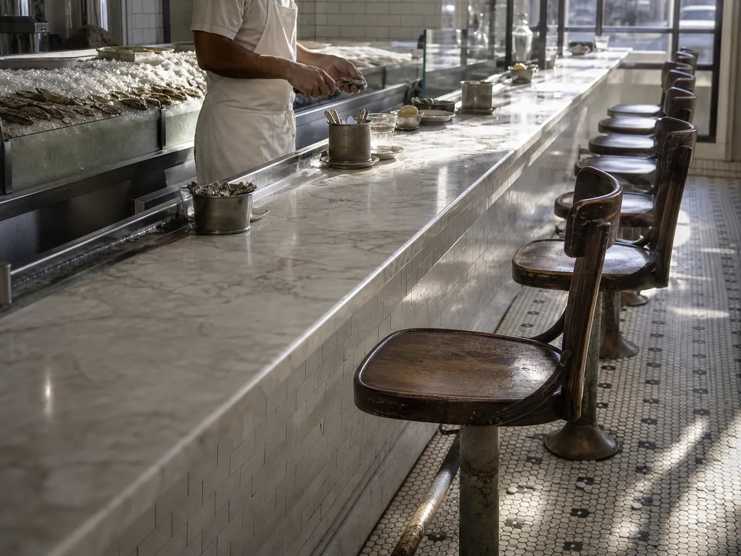 Long white marble oyster counter with a row of worn wooden swivel stools and trays of shellfish on ice in a century-old San Francisco seafood shop