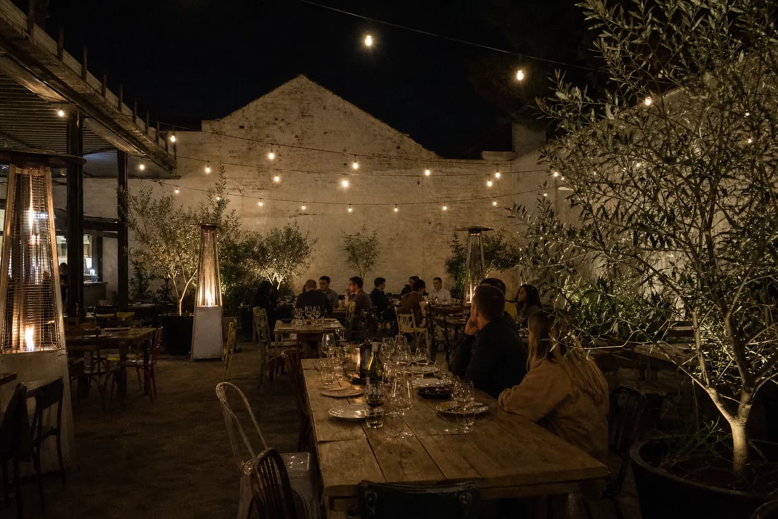 Night view of a partially covered outdoor dining courtyard with long communal tables, strings of small warm bulbs overhead and patio heaters