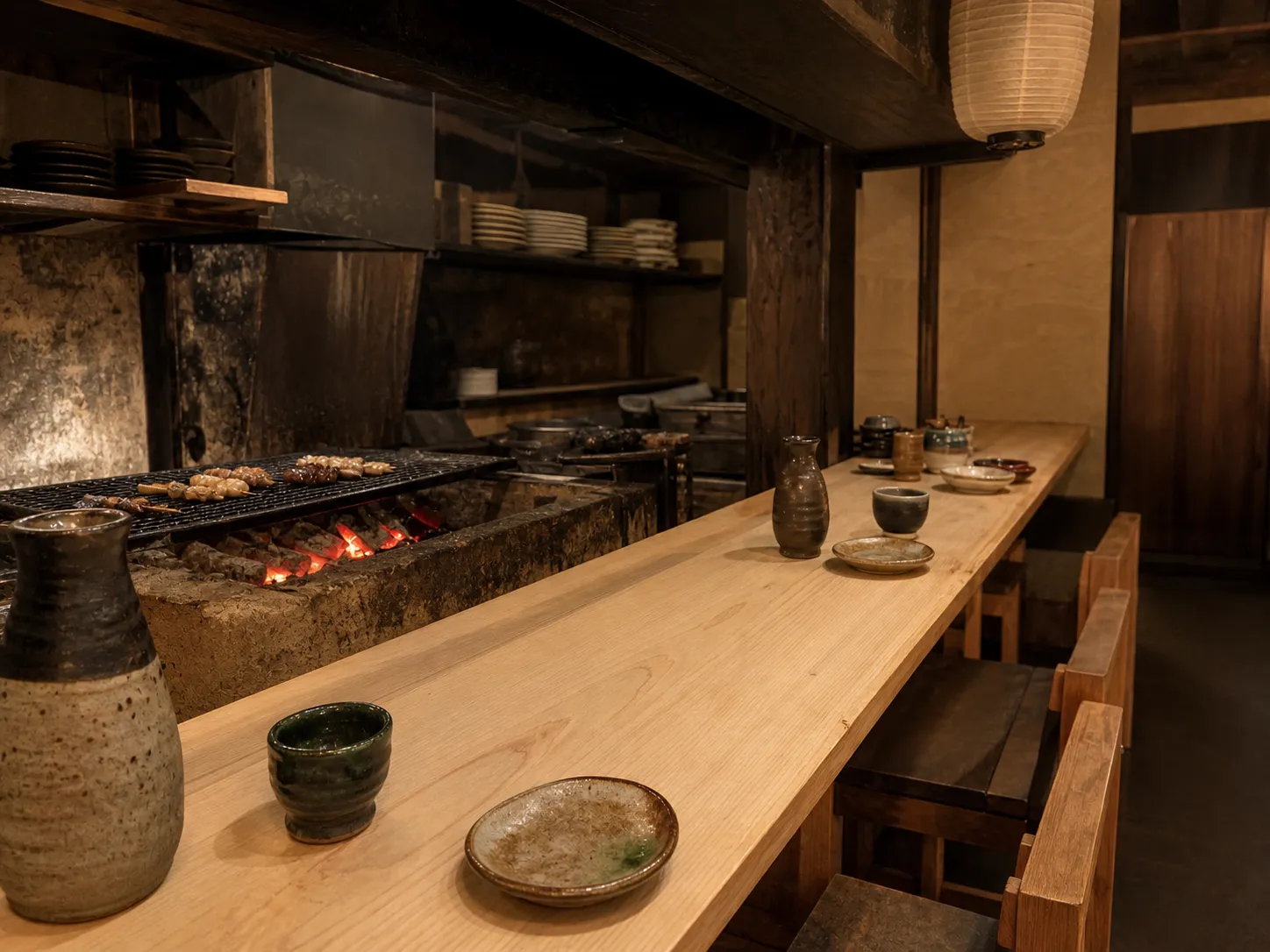 Interior of a small Japanese izakaya with a long pale wood counter, wooden stools, ceramic sake cups and a glowing charcoal grill behind the counter under warm dim light