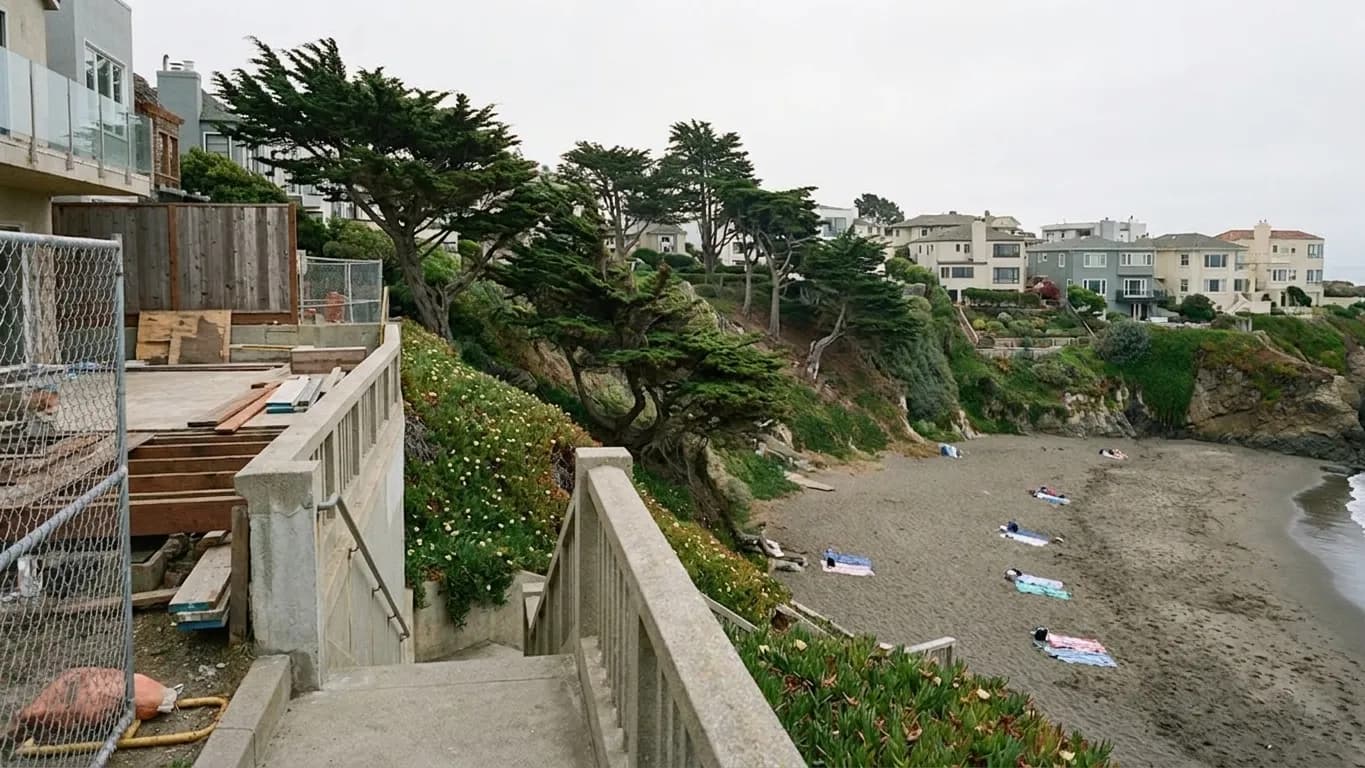 A small crescent cove beach in San Francisco's Sea Cliff seen from the stair landing above, with a concrete stairway and railing descending the bluff, construction fencing on the upper terrace, and a compact strip of sand below under flat grey coastal light