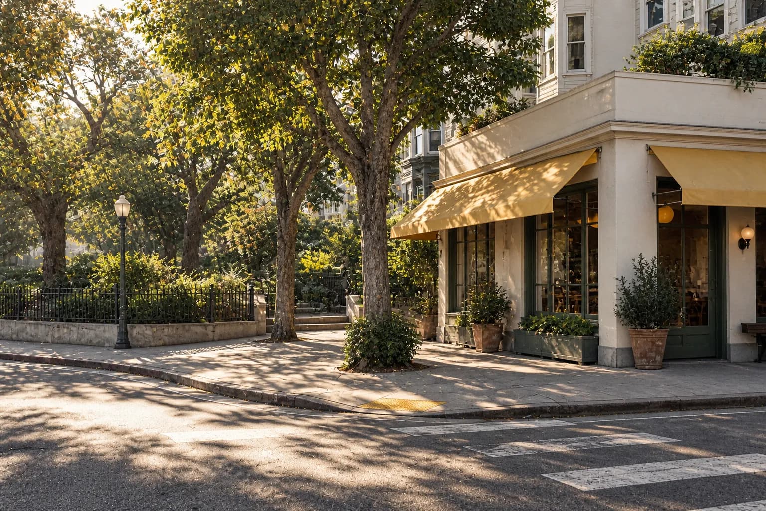 Wide morning view of a low-rise restaurant frontage beside a leafy city park, with a corner street intersection, pale building walls, shaded pavement and warm sunlight across the public square.