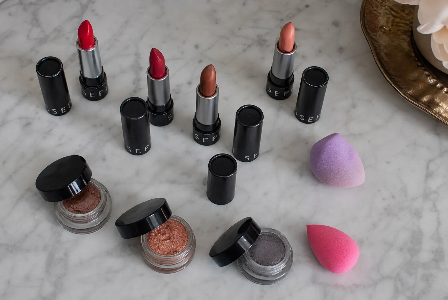A minimalist tabletop flat lay of Sephora brand lipsticks, cream blushes, and makeup brushes arranged on a clean white surface with soft shadows.