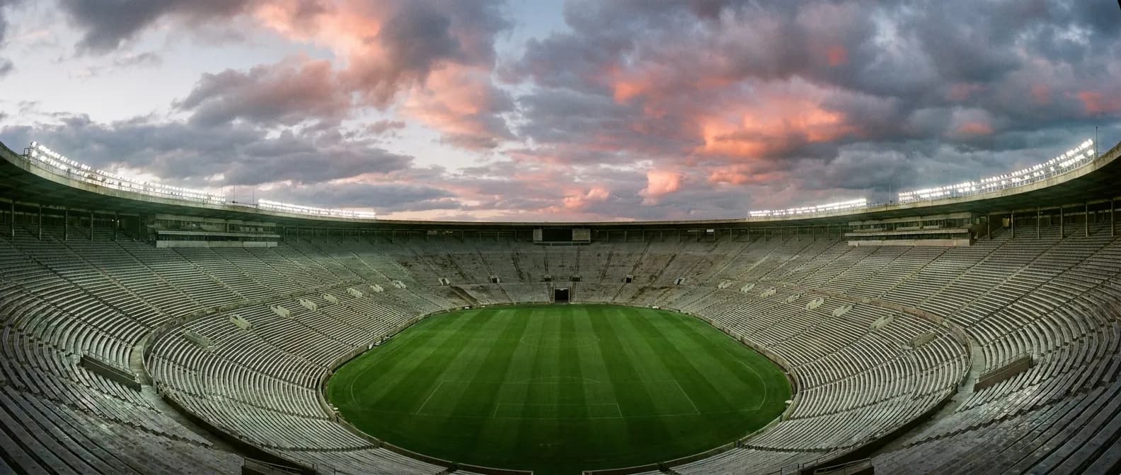 A vast oval stadium bowl glowing under lights at dusk with a dramatic sky overhead