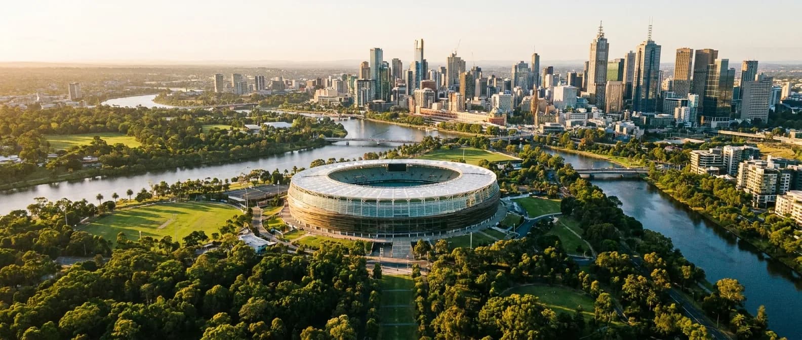 Aerial view of an oval sports ground amid green parkland beside a city skyline at golden hour