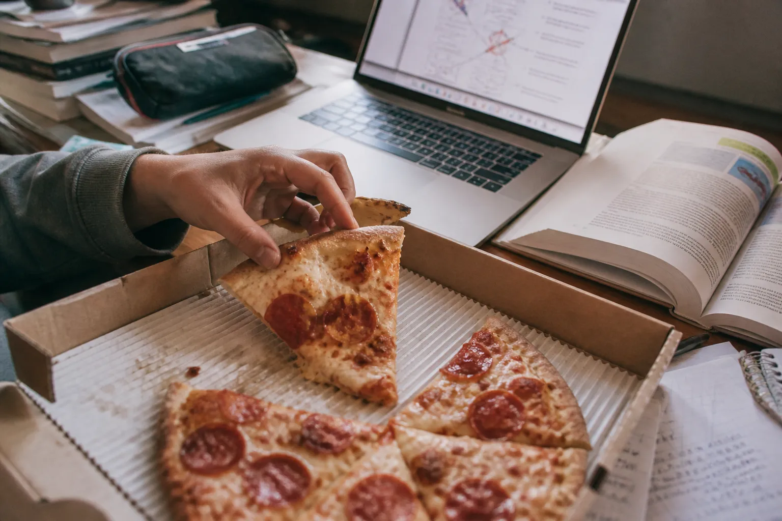 A slice of pepperoni pizza being pulled from a box on a student's desk with study materials.