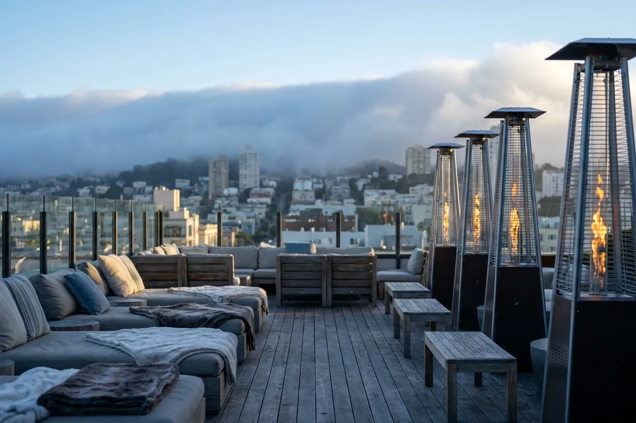 A San Francisco rooftop terrace in the late afternoon with timber decking, patio heaters and fog beginning to spill over the hills behind the skyline