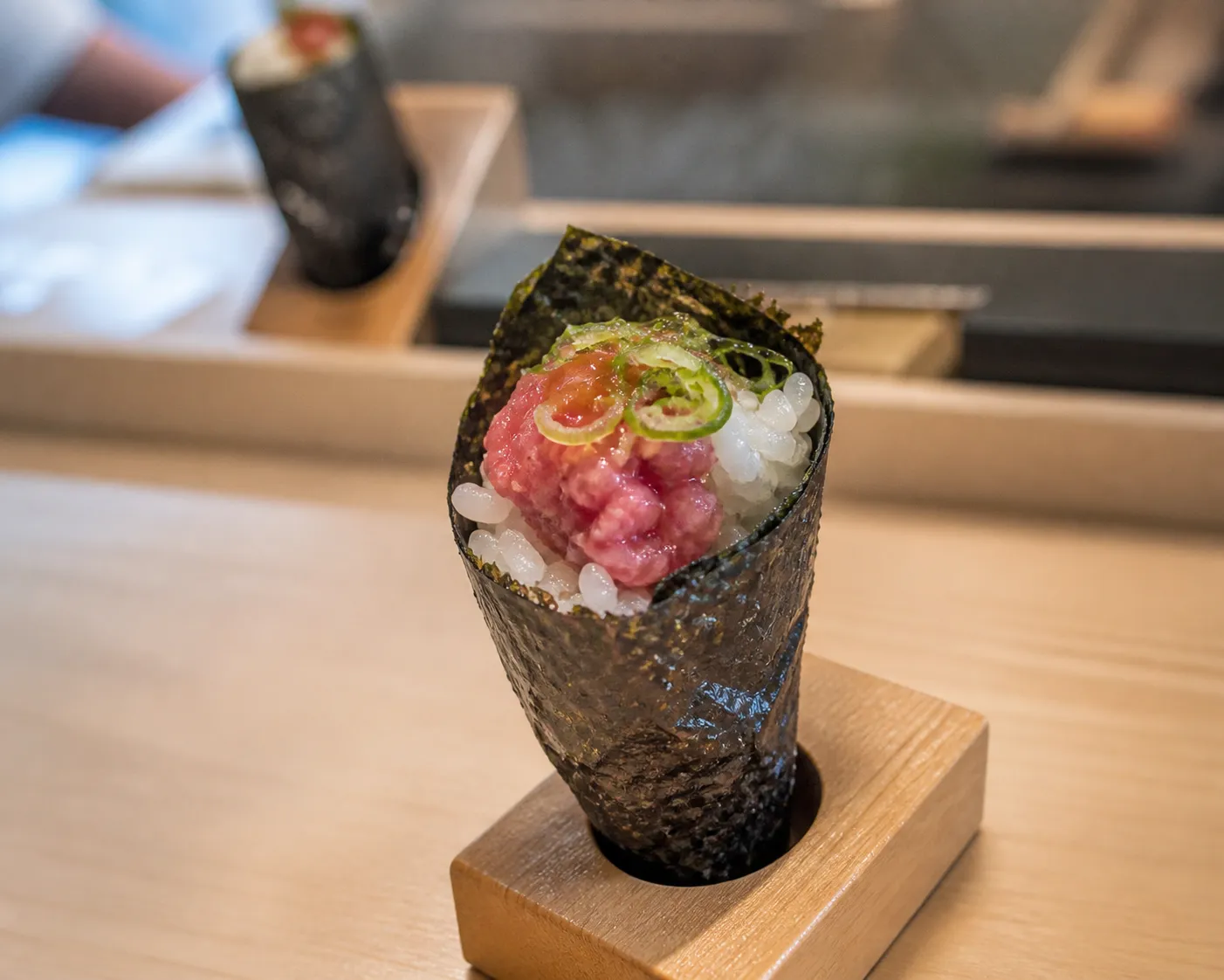Close-up of a crisp nori hand roll standing in a wooden holder on a light wood sushi counter, filled with rice, raw fish and green onion
