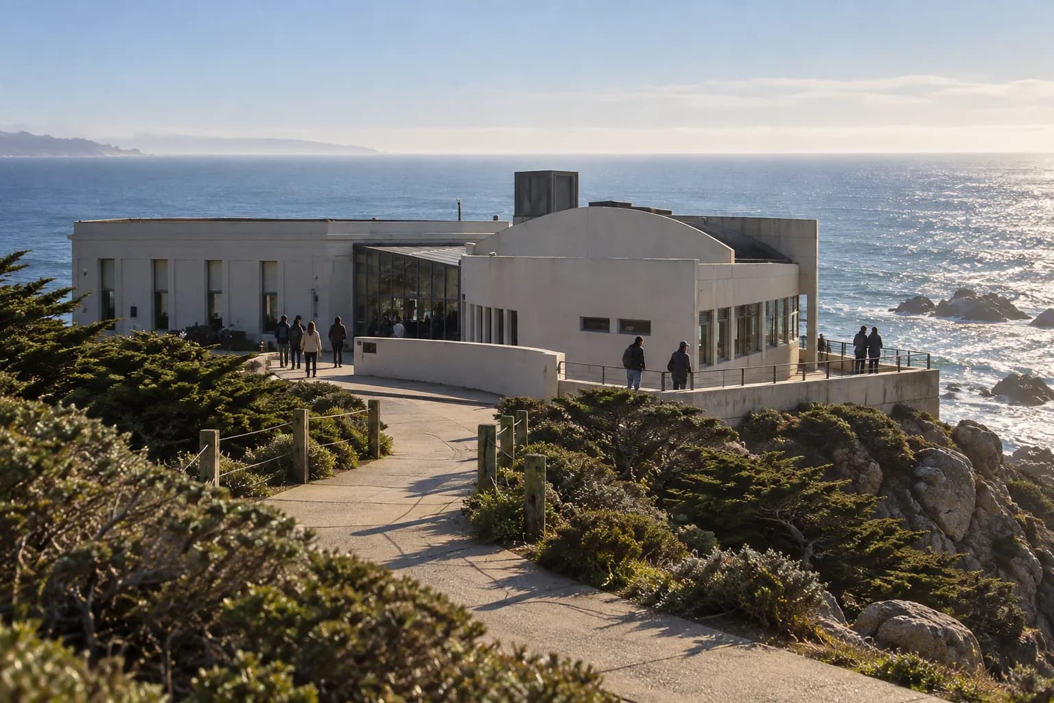A small camera-shaped building painted pale on a windswept cliff above the Pacific, the ocean flat and bright behind it
