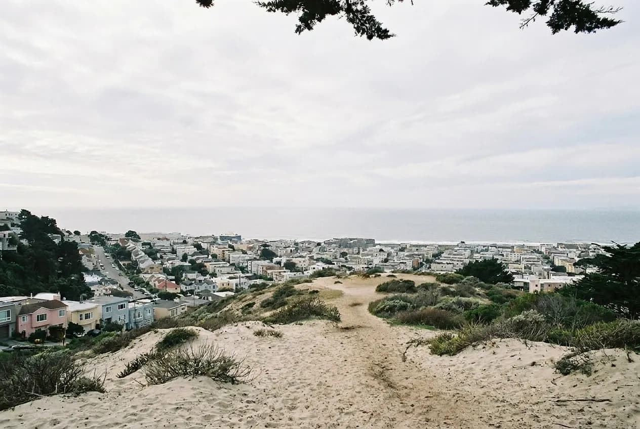 A bare sandy hilltop with low scrub and a wide city view beyond