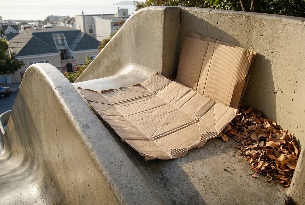 A flattened sheet of cardboard resting on the concrete lip at the top of a slide, with the city rooftops below