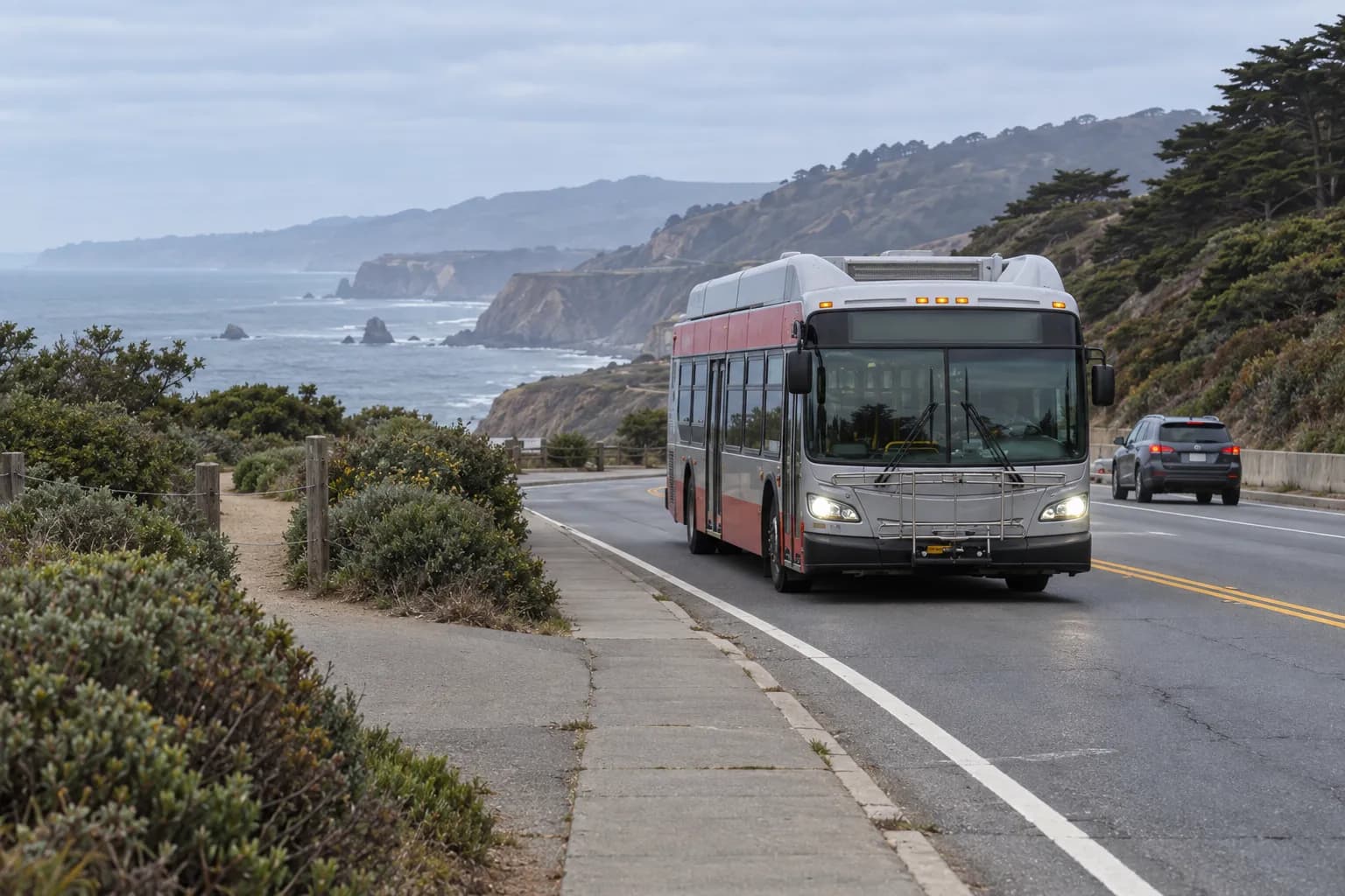 A city bus travels along a coastal San Francisco road near a trail entrance, with scrub, pavement and distant ocean cliffs framing the route towards a historic shoreline site.