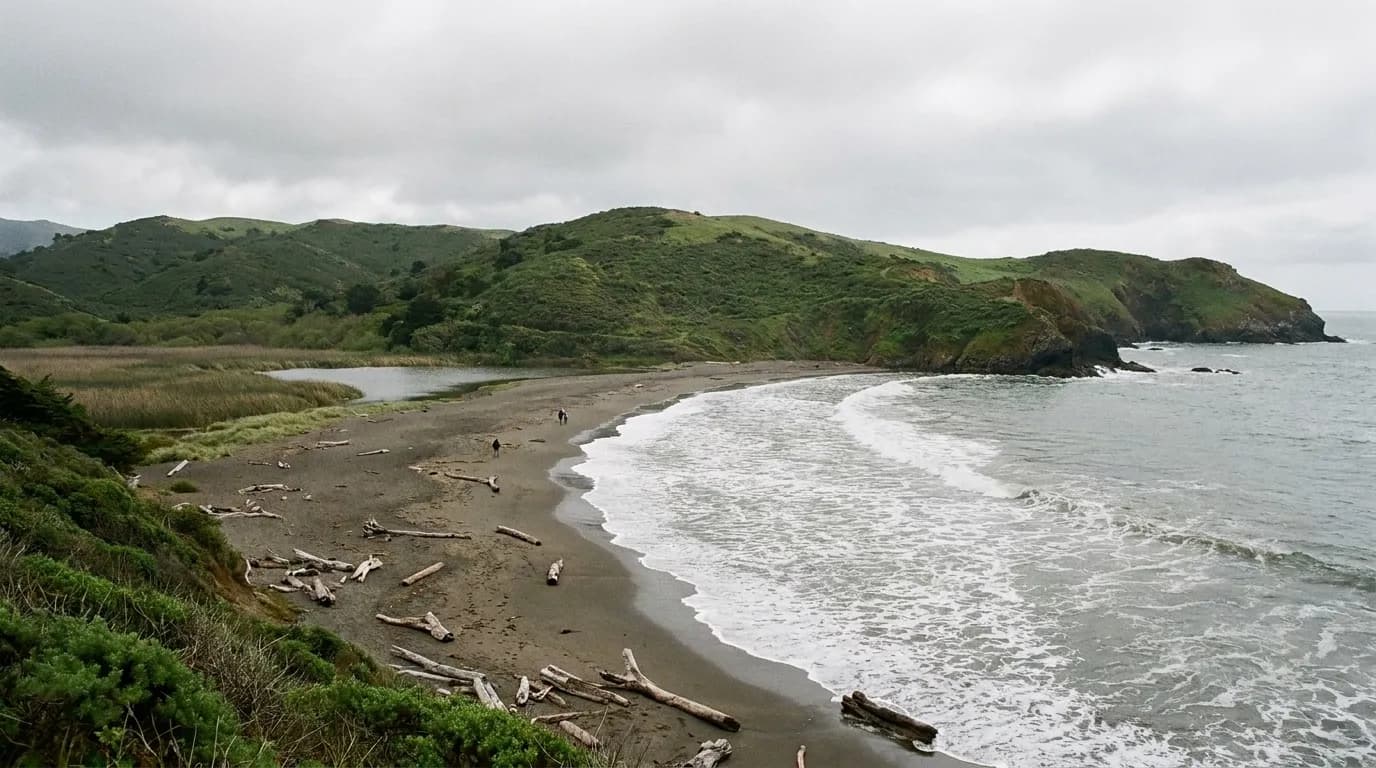 A small crescent beach in Marin County backed by green hills, with a shallow lagoon behind the sand under grey cloud.