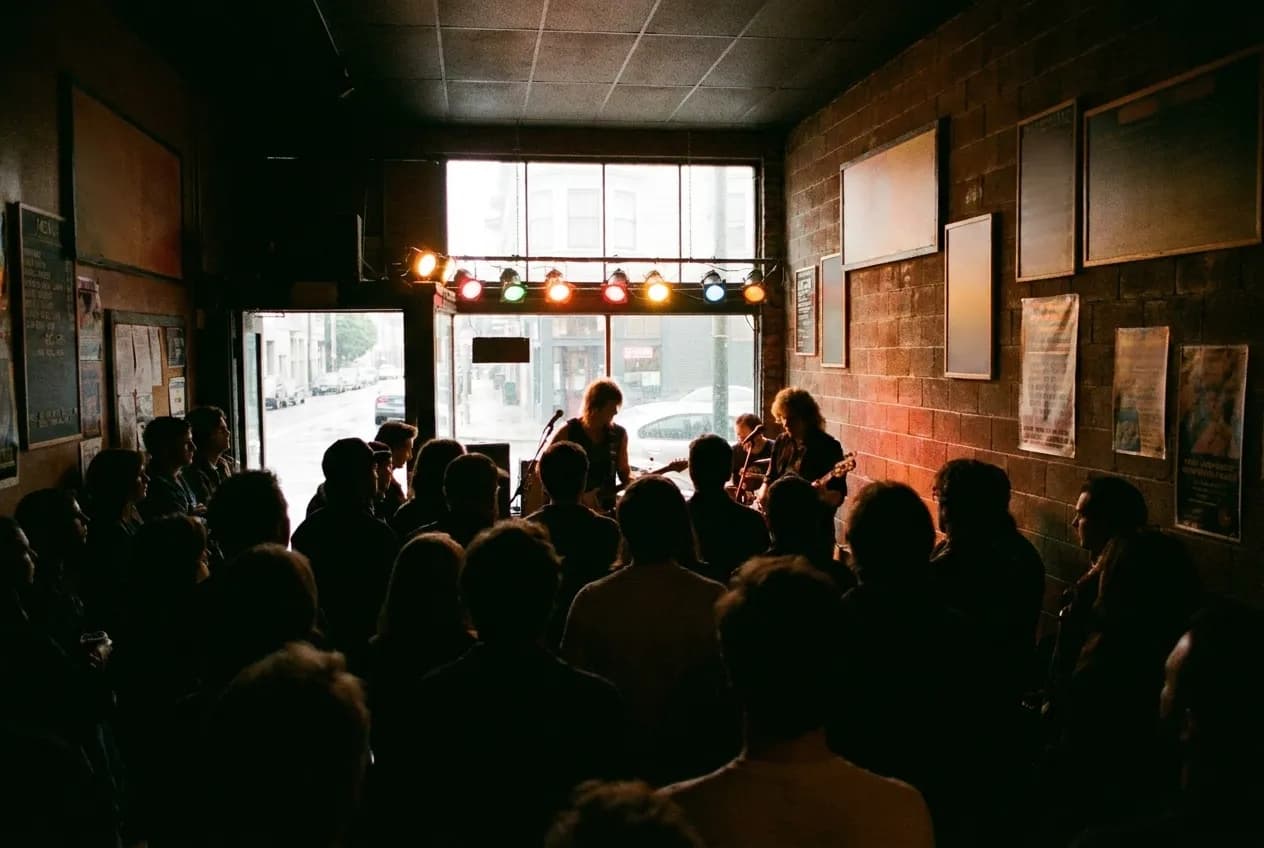 A small venue seen from the back of the room, stage lit, heads in silhouette