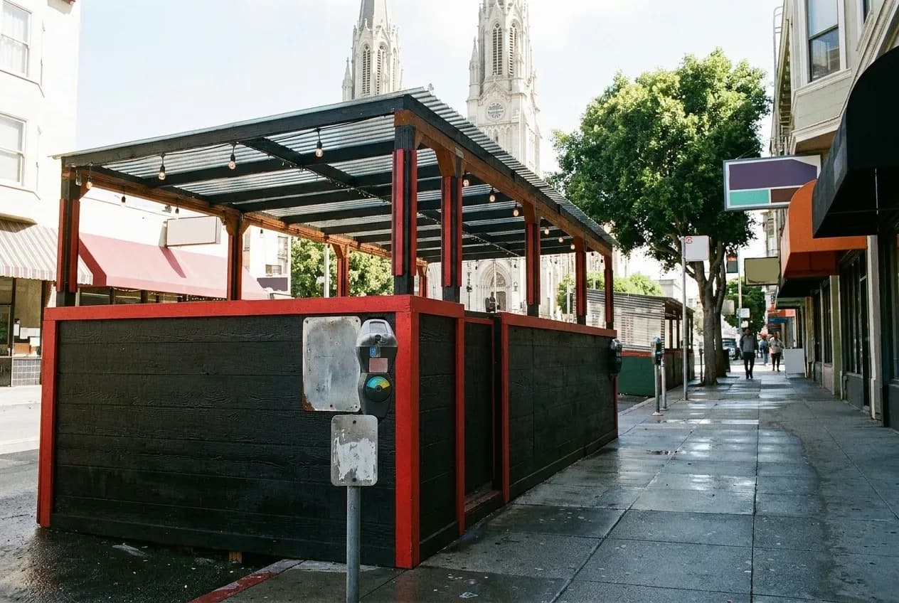 A covered parklet on a wide pavement, church towers rising behind it down the block