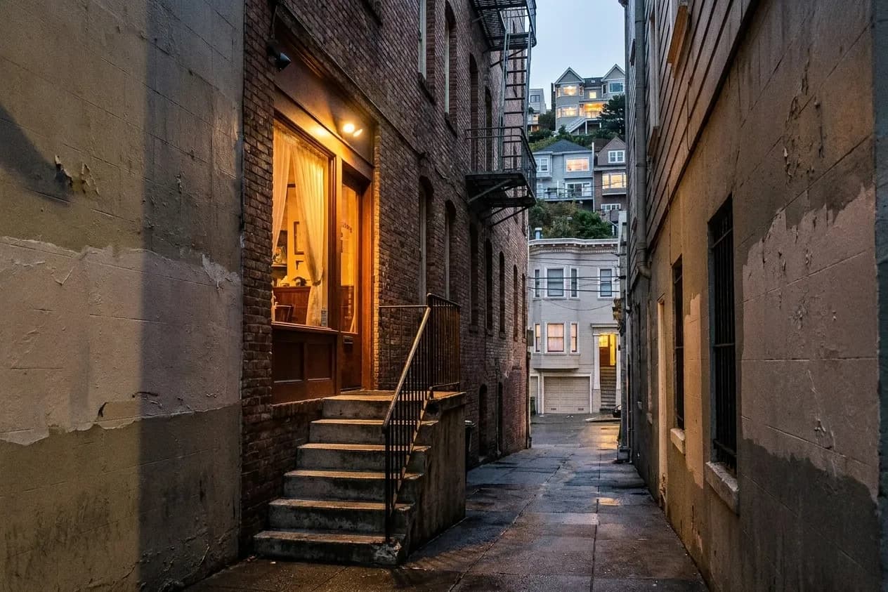 A narrow San Francisco side street at dusk with concrete steps up to a recessed restaurant door, warm light from a curtained window and a fire escape zig-zagging above against the lit hill beyond
