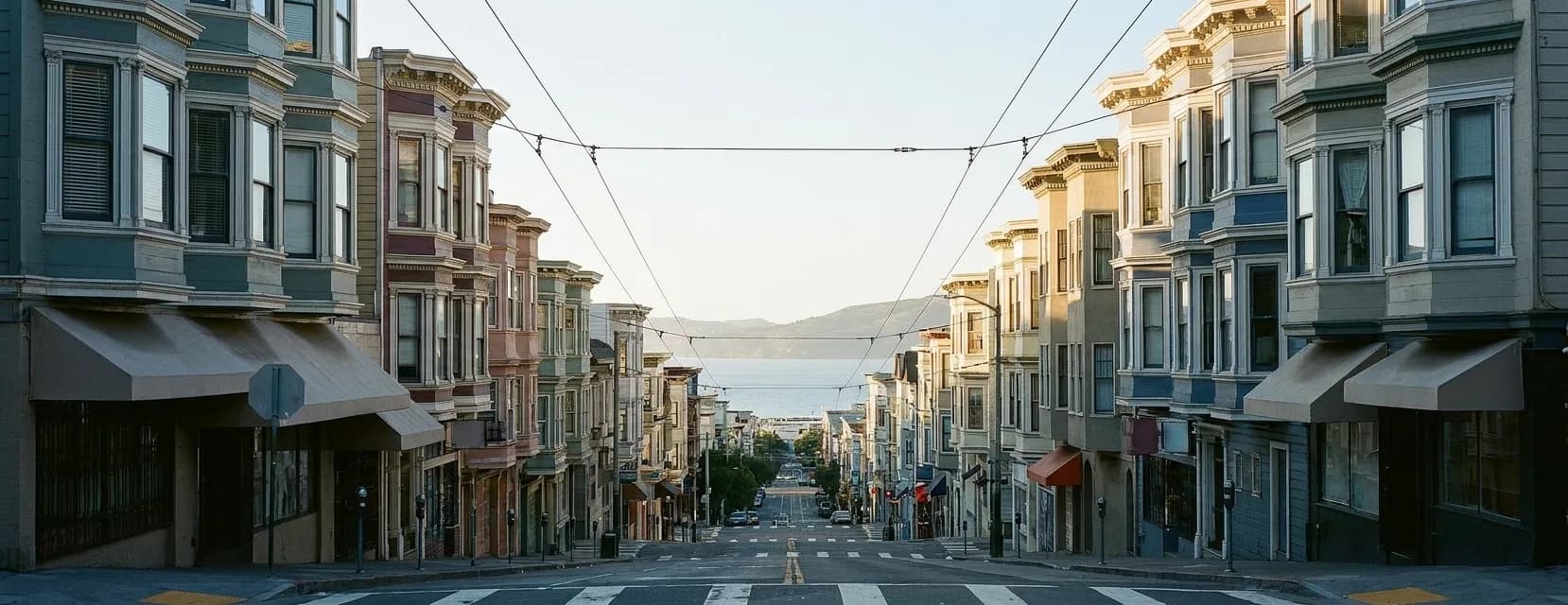 A San Francisco commercial street in late afternoon seen from a slight rise, with bay-windowed storefronts stepping down the hill, wires strung overhead and a wedge of pale bay water at the end of the block