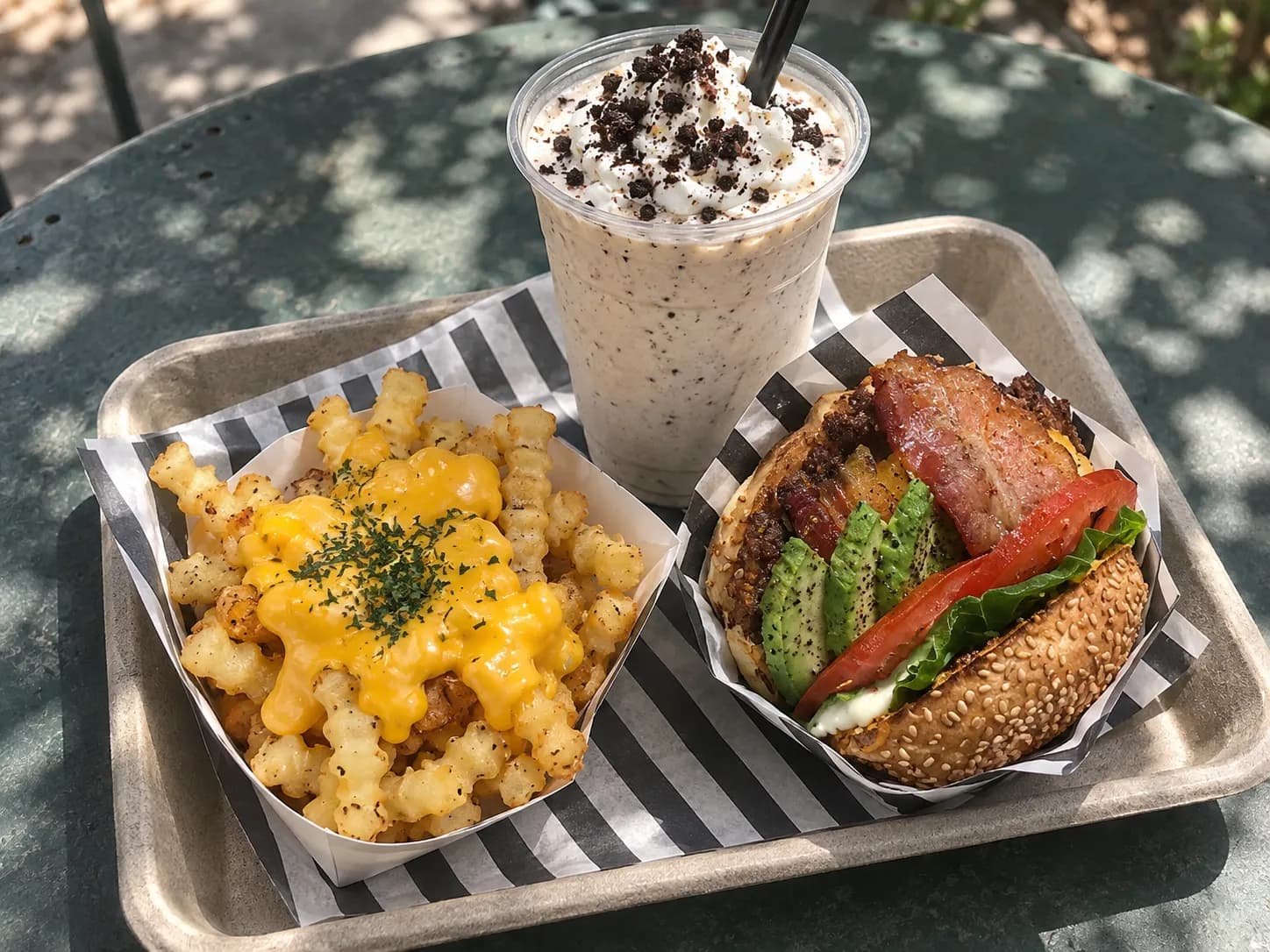 Small toasted-bun cheeseburger beside a paper cup of crinkle-cut fries and a thick milkshake on a tray with a paper liner in dappled tree shade