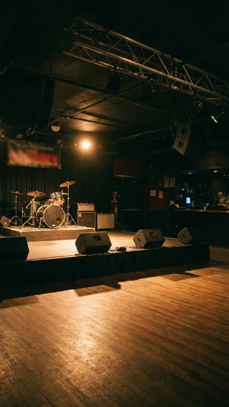 A small dark music venue before doors with a low stage, a drum kit under a single spotlight and empty floor space in front
