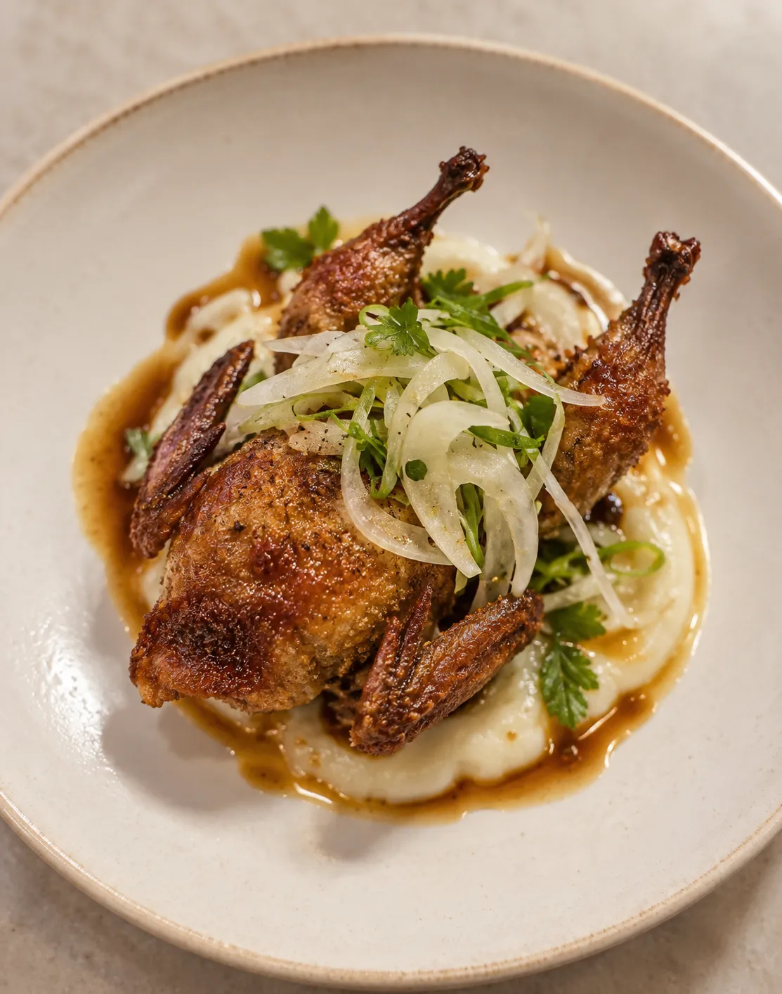 Small crisp fried quail glazed dark golden on a shallow dish, resting on pale onion-cream with a light salad of shaved onion on top