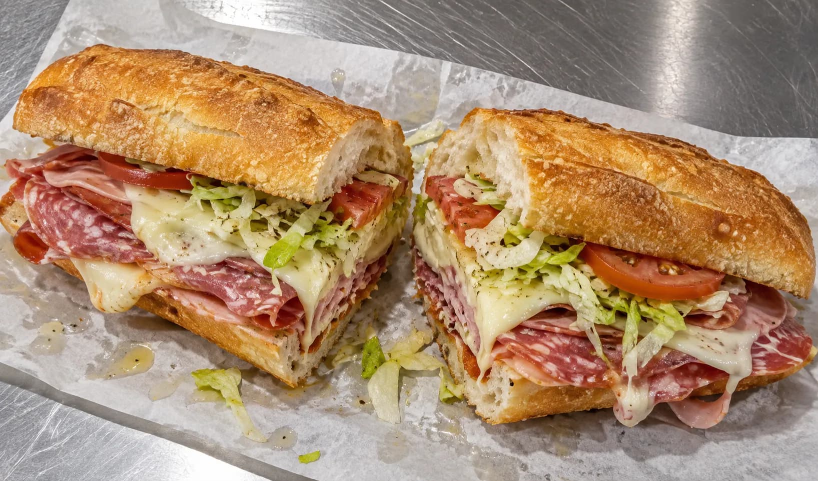 Hot Italian sub on a long roll cut in half on greaseproof paper, with layered cured meats, melted cheese, lettuce and tomato at the cut faces
