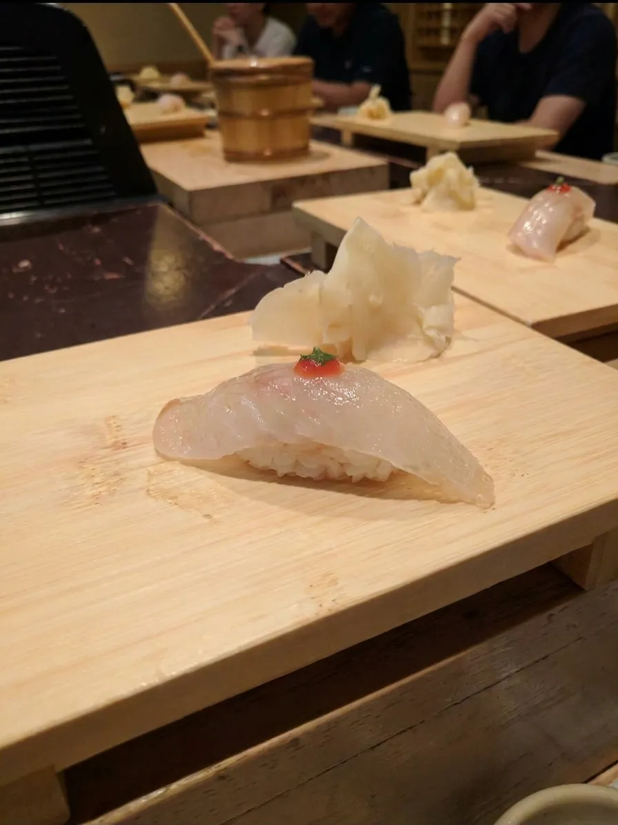 Single piece of white fish nigiri on a pale wooden serving board with pickled ginger, shot tabletop-level at a New York sushi counter
