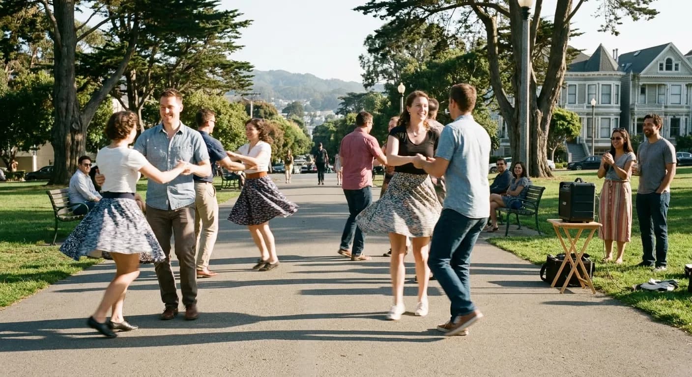 People swing dancing outdoors on a paved park path on a sunny weekend, several couples mid-turn with a portable speaker on a folding table at the edge and onlookers around the perimeter