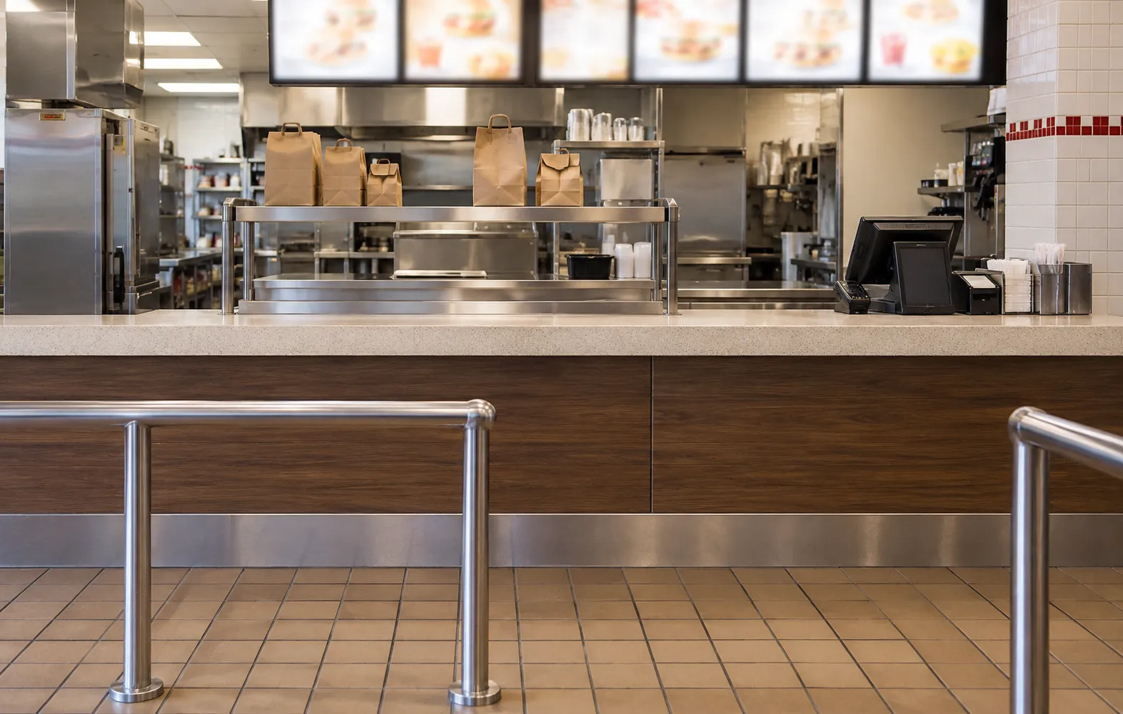 Fast-food service counter with a stainless pickup shelf holding paper bags, a register position to one side and a queue rail in the foreground
