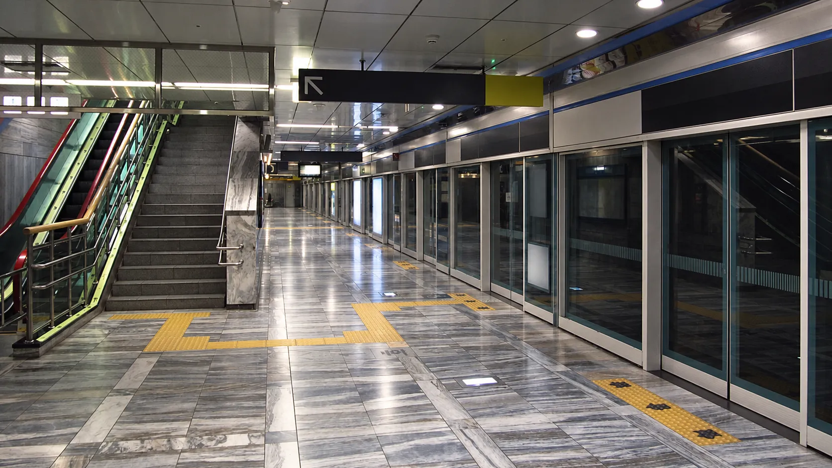 Underground transit station concourse with escalators rising toward a lit mezzanine and a tiled platform edge with a yellow safety strip