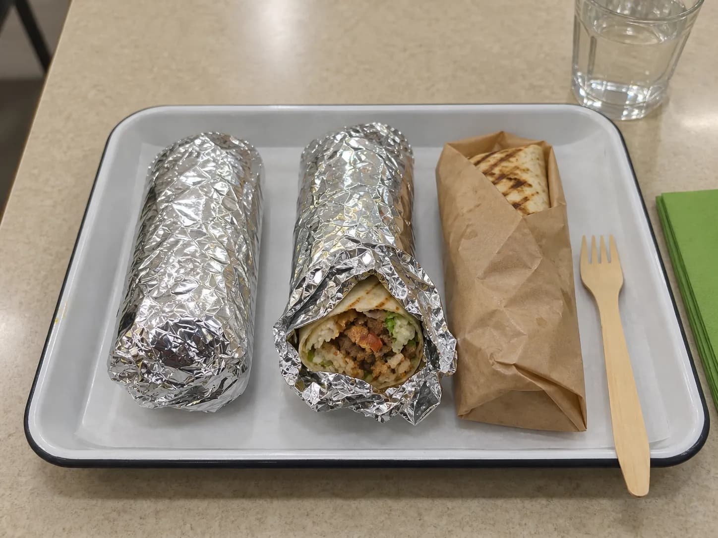 Two foil-wrapped items and a paper-wrapped grilled item on a moulded plastic tray with a paper liner, one partly unwrapped showing the filling