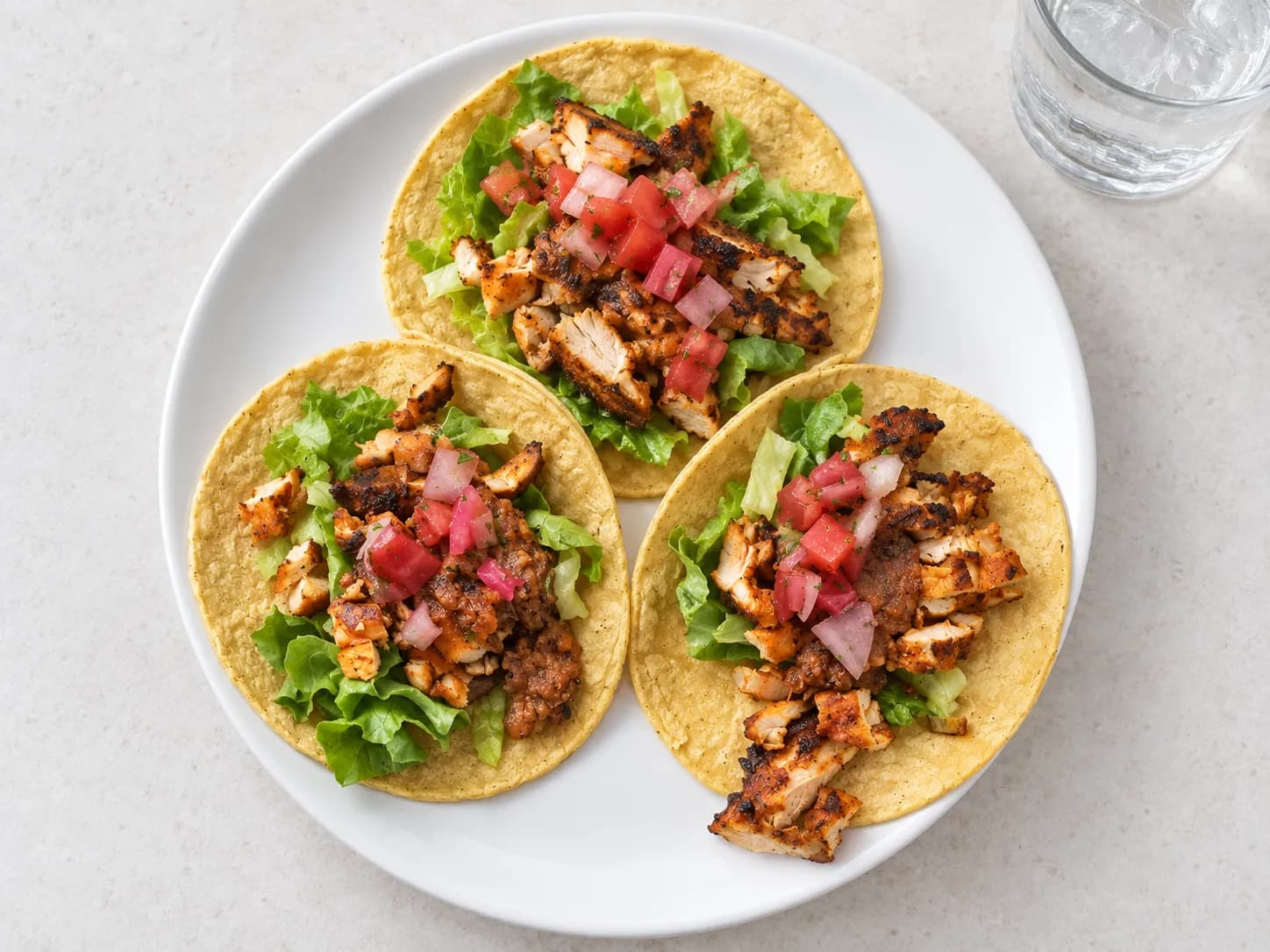 Three soft tacos filled with grilled sliced chicken, shredded lettuce and diced tomato on a plain white plate beside a glass of water