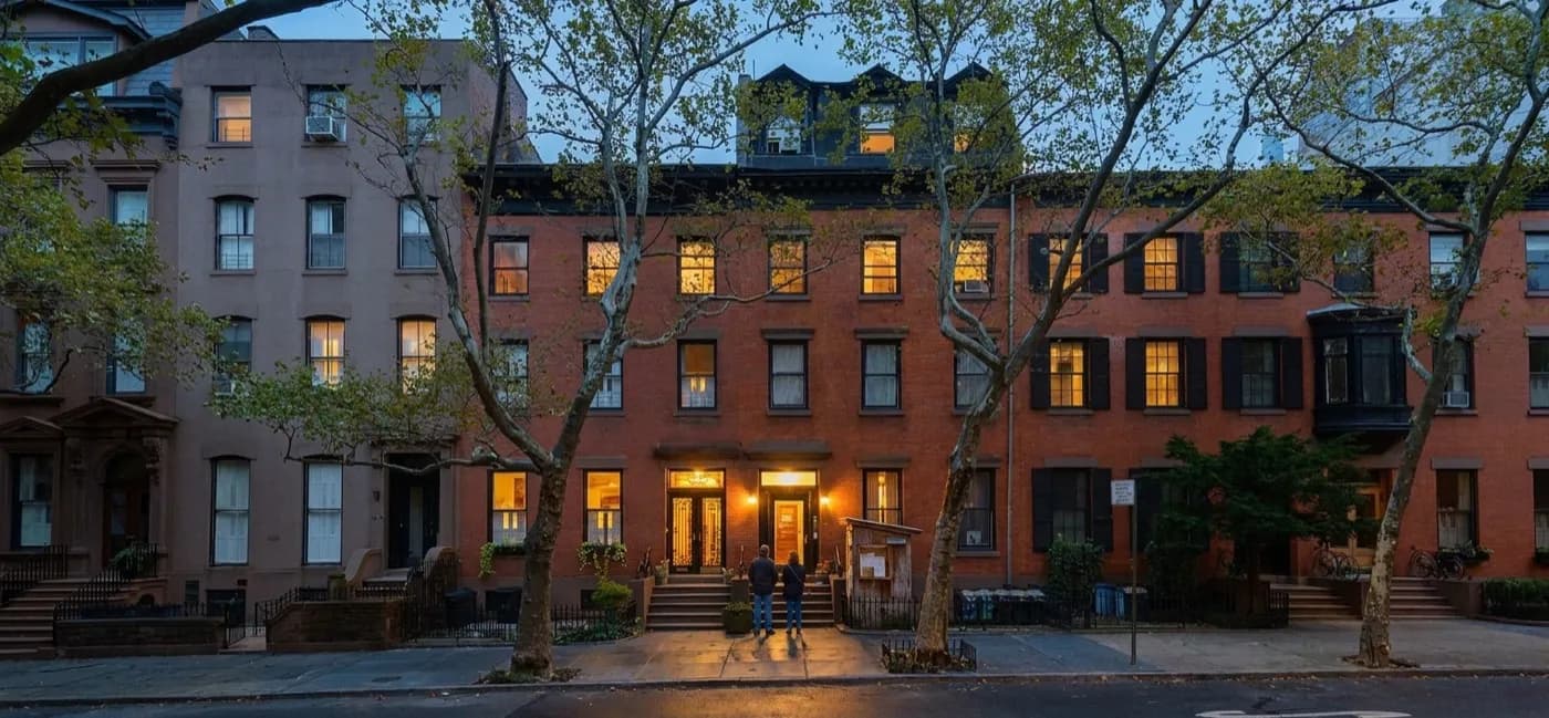 A lit restaurant doorway on a quiet Brooklyn side street at early evening with a handwritten list on a stand and two people waiting on the stoop