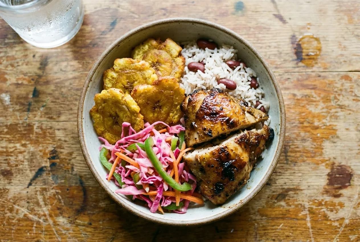 An overhead view of a Caribbean jerk chicken bowl with plantains, pikliz, and rice on a wooden table.