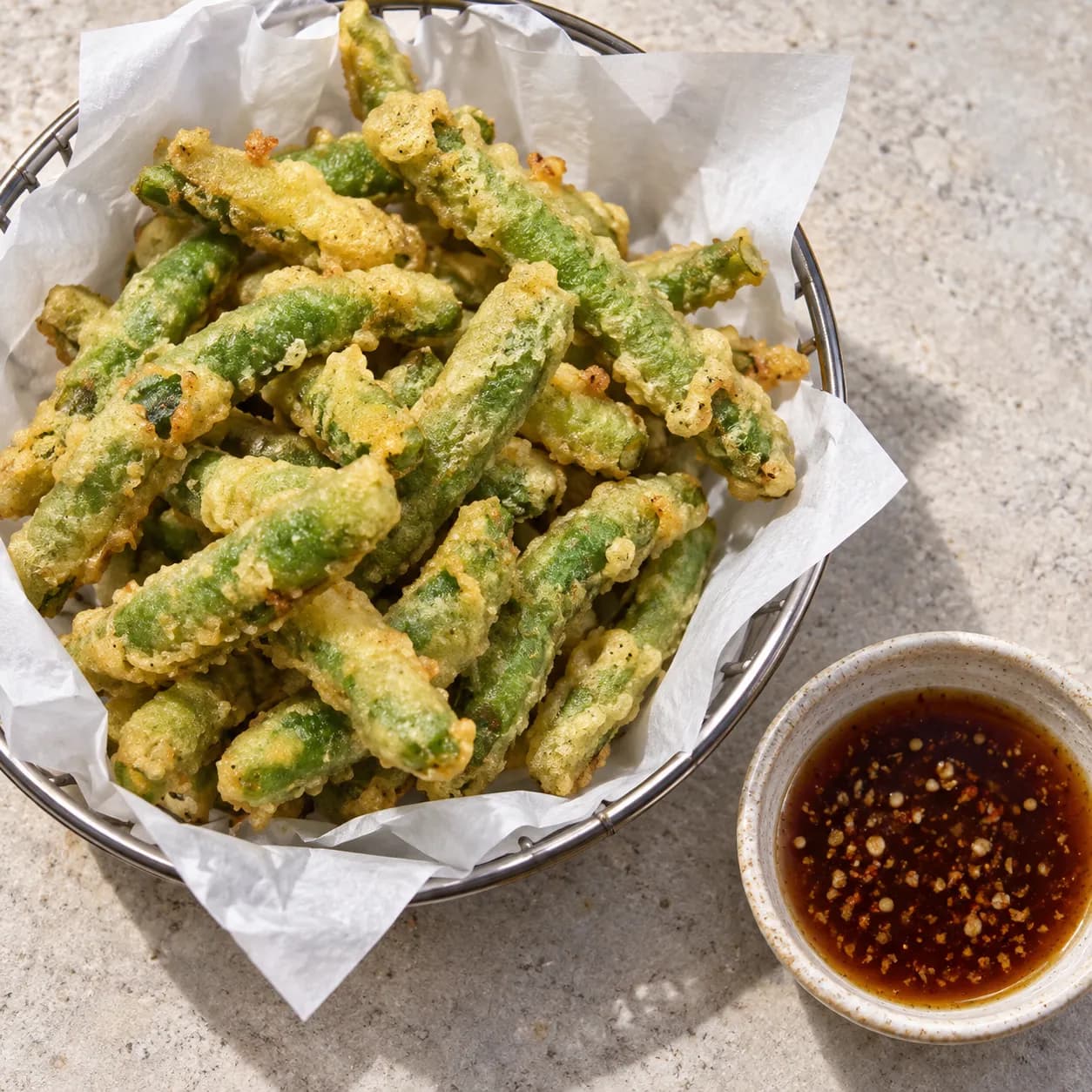 Battered tempura green beans piled in a paper-lined basket with a light crisp blistered coating, beside a pot of dipping sauce