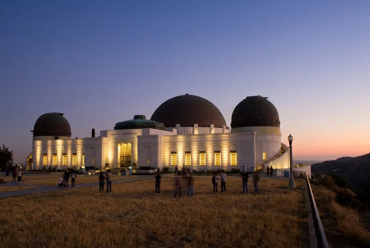 Griffith Observatory floodlit on its hilltop lawn at dusk with visitors on the grass