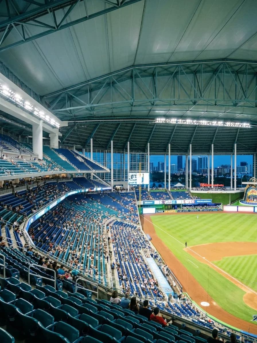 The interior of a modern Miami ballpark in the evening with the retractable roof closed, floodlit green infield below half-filled teal seat banks and the city skyline visible through the glass outfield wall