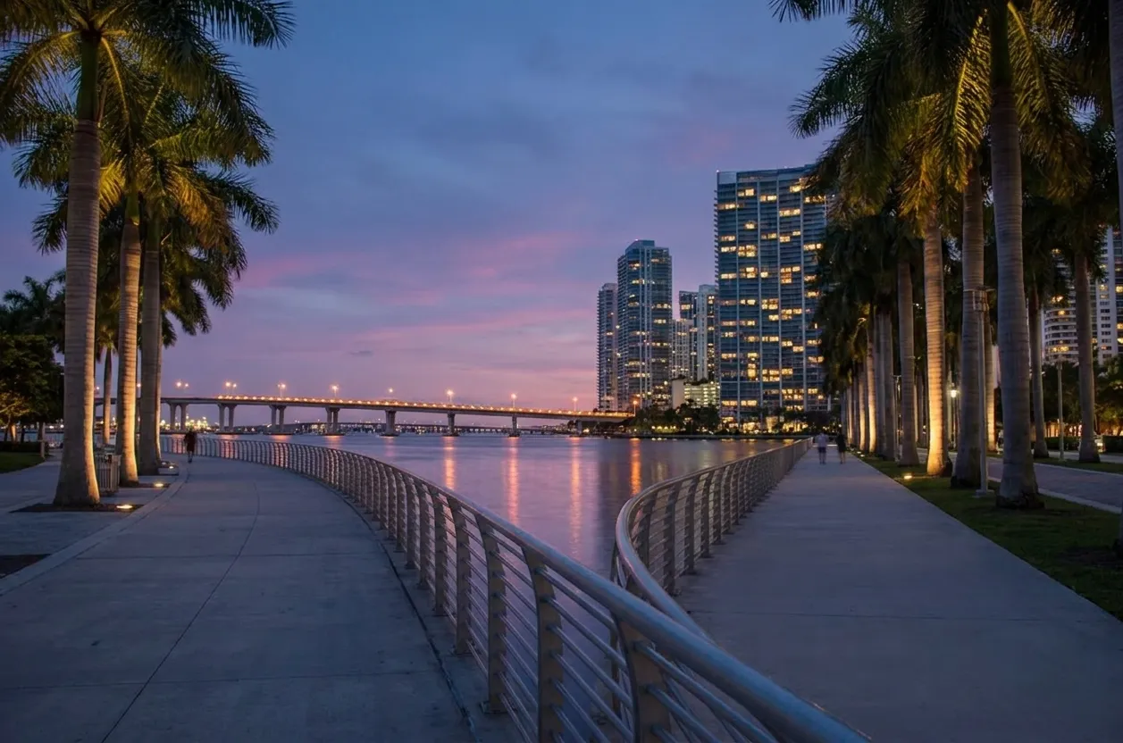 Fifteen minutes after sunset on the downtown Miami baywalk, a railed promenade curving along Biscayne Bay with uplit palms, a lit causeway bridge and glass towers reflecting pink and violet on the water