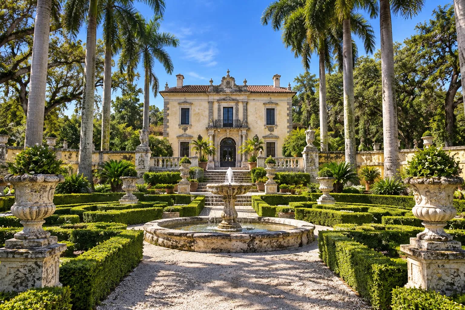 A formal garden of clipped hedges and stone urns leading toward a large Italianate villa under palms