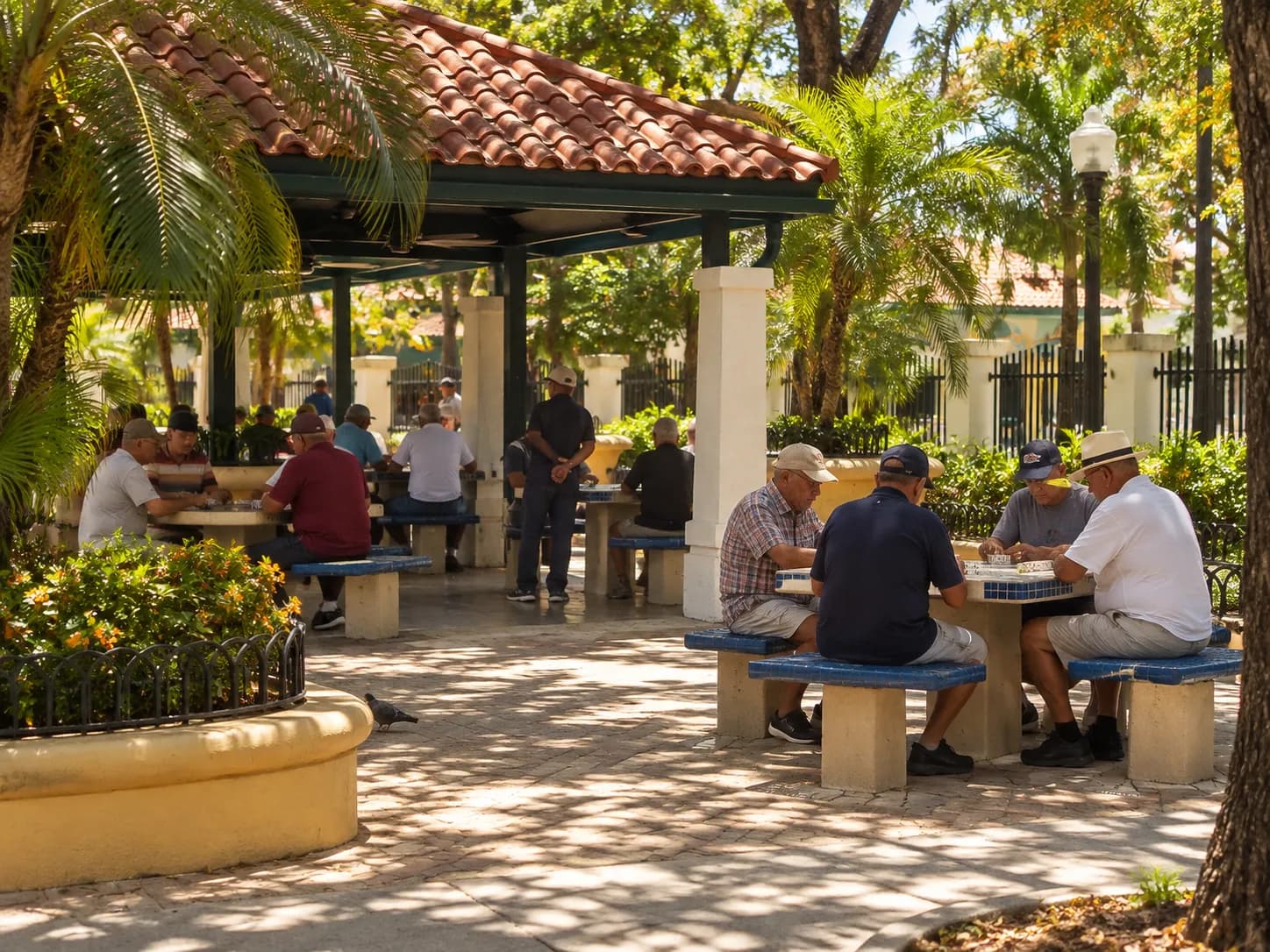 Older men playing dominoes at shaded outdoor tables in a small park, with palms and a tiled roof above