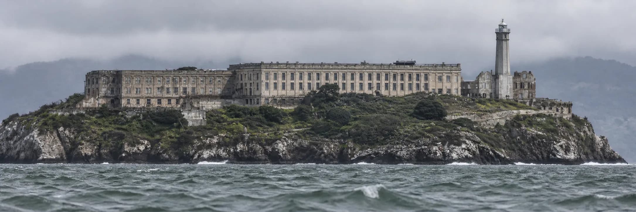A rocky island with institutional buildings and a lighthouse seen across open water