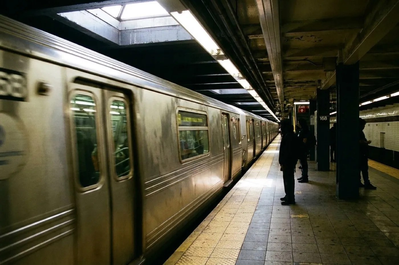 A subway platform with a train arriving, sunlight falling through a station skylight onto the tiled floor and waiting passengers