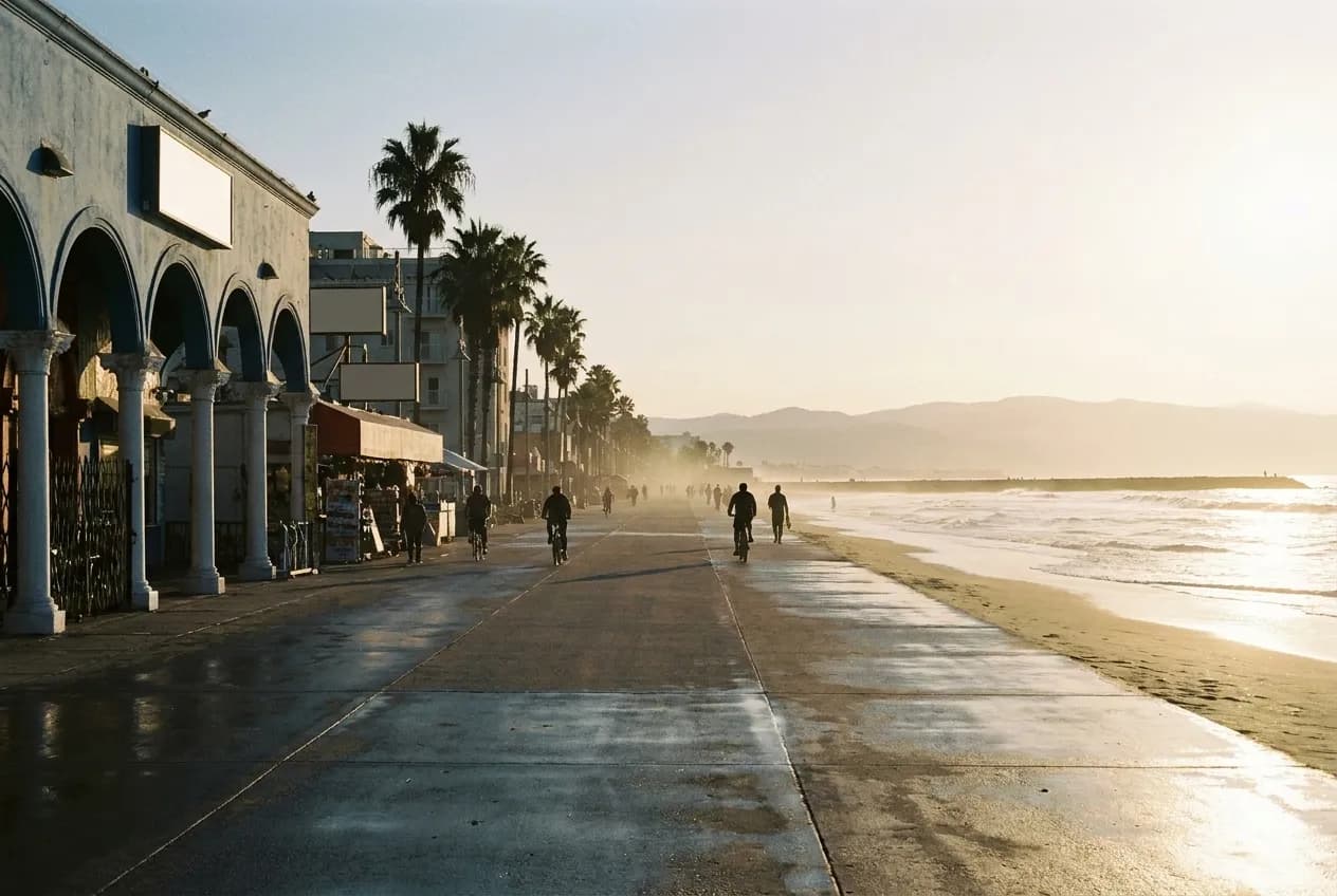 The Venice Beach Boardwalk stretching along the Pacific Ocean with vendor stalls, palm trees, and cyclists in late afternoon light.