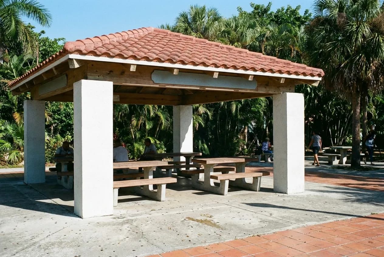 An open-sided shelter with a tiled roof over fixed tables and benches in a small urban park, dense planting behind and hard midday shadow underneath