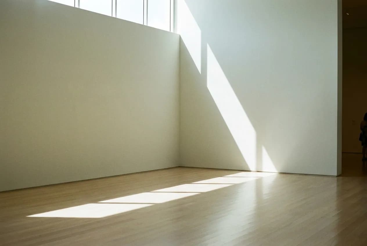 A tall gallery space with pale walls and polished floor, daylight from a high window falling across an empty corner of the room