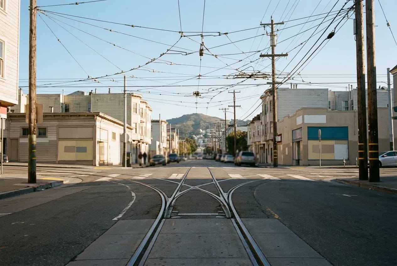 Rails set into a city street with overhead wires converging above, low buildings on both sides and a hill rising in the background under soft light