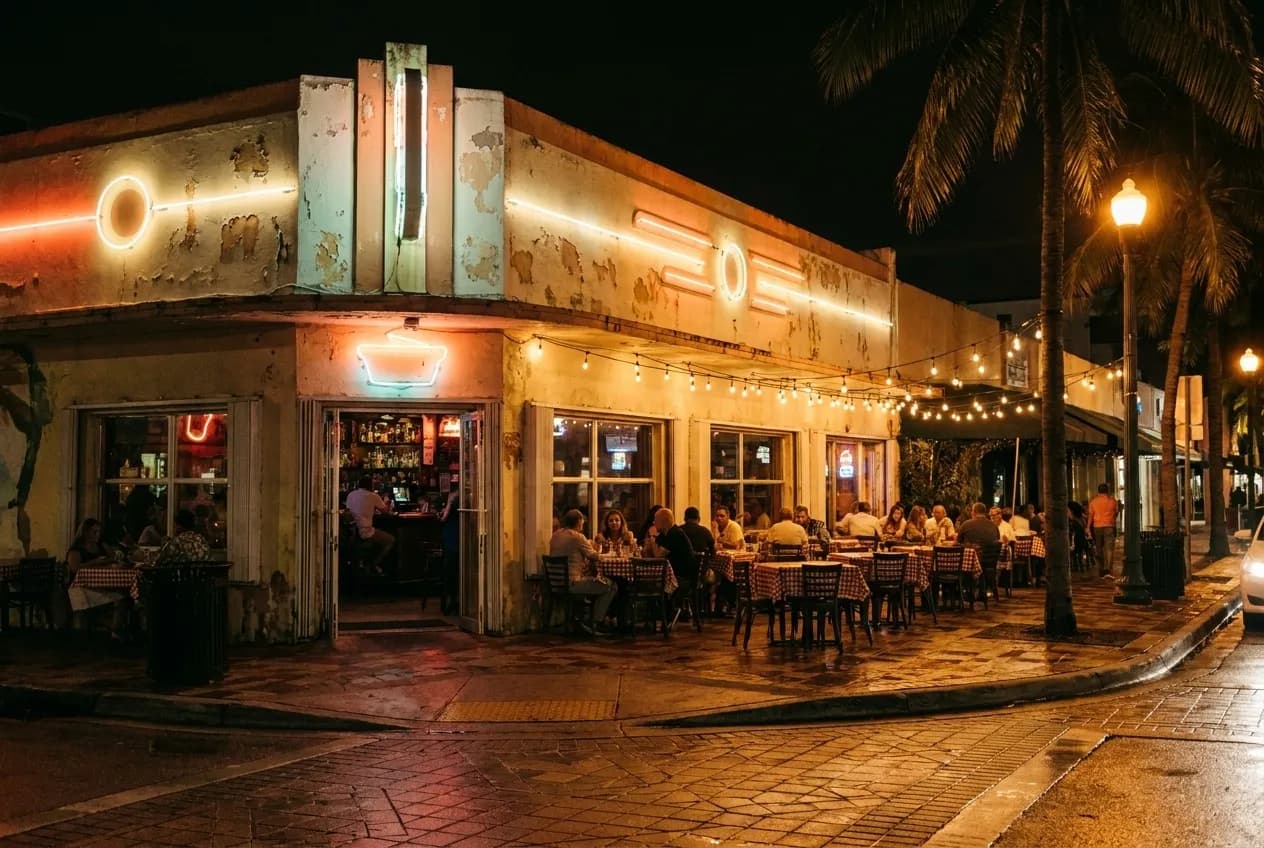 Neon bar signage and art-deco lettering illuminate a corner storefront on Calle Ocho at night, with café tables and string lights spilling onto the tiled sidewalk.